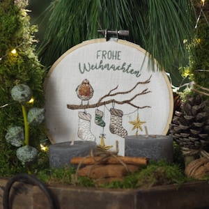 May include: A white embroidery hoop with the text "Frohe Weihnachten" and an embroidered image of a robin perched on a branch with three stockings hanging from it. The hoop is surrounded by greenery, cinnamon sticks, and gray candles.