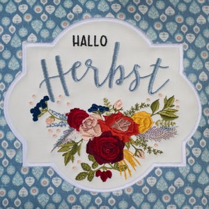 May include: A white embroidered patch with a floral design and the text "Hallo Herbst" in blue lettering.