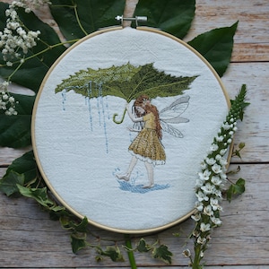 May include: Embroidered hoop art featuring a fairy holding a green leaf umbrella, with blue raindrops falling. The fairy is wearing a yellow dress and has brown hair and wings.