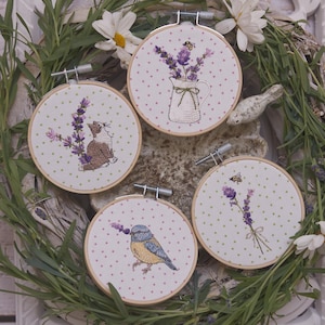 以下が含まれることがあります： 花と動物のデザインが施された4つの刺繍フープ。1つはラベンダーと猫、もう1つはラベンダーと鳥、3つ目はラベンダーの花瓶、最後はミツバチとラベンダーの束です。フープは緑の背景に配置されています。