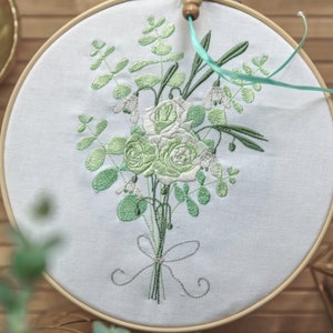 May include: A white embroidery hoop with a green floral design. The design features roses, leaves, and small white flowers. The hoop is tied with a white ribbon.