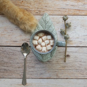 May include: A cup of hot chocolate with marshmallows on a gray leaf-shaped coaster. A silver spoon and a sprig of dried flowers are on a wooden surface.