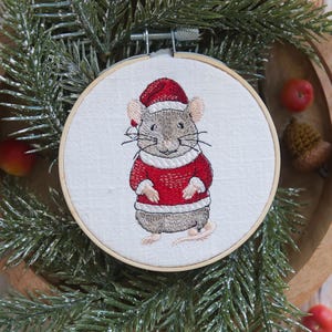 Può includere: Ornamento natalizio ricamato con un topo grigio che indossa un cappello e un maglione rossi di Babbo Natale. Il topo è ricamato su tessuto bianco, incorniciato da un telaio da ricamo in legno. Decorazione festiva natalizia.