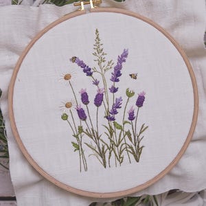 Op de afbeelding: Geborduurde bloemenhoepelkunst met lavendel, madeliefjes en groen. Het ontwerp bevat twee bijen en is geplaatst tegen een witte stoffen achtergrond. De houten hoepel heeft een natuurlijke kleur.