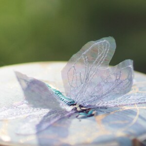 Könnte beinhalten: Ein zarter, hellblauer Schmetterling, der auf einem weißen Stoffhintergrund gestickt ist. Die Flügel des Schmetterlings sind durchscheinend und haben ein zartes, gesticktes Muster.