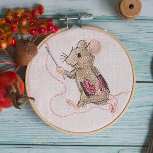 May include: Embroidered artwork in a wooden hoop features a mouse holding a needle and thread. The mouse is brown and tan with pink accents. The background is white fabric. The hoop is surrounded by fall foliage.