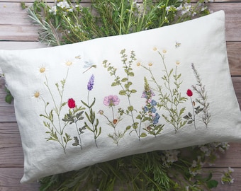 Puede incluir: Una almohada blanca con flores silvestres bordadas en tonos de rosa, morado, azul, rojo y verde. La almohada está sobre una superficie de madera con follaje verde.