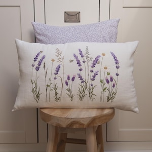 May include: A rectangular white pillow with an embroidered lavender and daisy design. The pillow is decorated with purple lavender flowers, white daisies, and green stems. A second pillow with a lavender floral pattern is behind it.