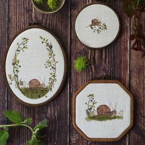May include: Three embroidered hoop art pieces featuring a snail design. The hoops are white with brown embroidery thread and are framed in wood. The first hoop has a snail in a green leafy design, the second hoop has a snail in a green leafy design with a blue berry pattern, and the third hoop has a snail in a green leafy design with a blue berry pattern.