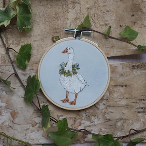 Könnte beinhalten: Gesticktes Kunstwerk einer weißen Gans mit einem Blumenkranz, in einem hölzernen Stickrahmen. Die Gans hat orangefarbene Füße und einen Schnabel. Der Hintergrund ist ein hellblauer Stoff. Grüner Efeu und ein Birkenrindenhintergrund umrahmen das Kunstwerk.