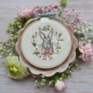 Pode incluir: Arte de bordado em aro com um coelho castanho com um laço rosa, rodeado de margaritas brancas sobre um fundo de linho branco.