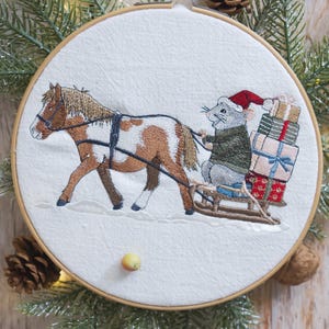 Può includere: Ricamo su telaio raffigurante un cavallo che traina una slitta con un topo con un cappello da Babbo Natale. La slitta è carica di regali confezionati. Il cavallo è marrone e bianco, e il topo è grigio. Lo sfondo è bianco, con aghi di pino e pigne.