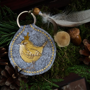 May include: A grey felt keyring with a gold embroidered bird wearing a crown. The keyring is attached to a silver key ring.