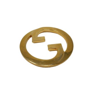 May include: A gold-toned metal belt buckle with a circular design featuring the interlocking 'G' logo of Gucci.