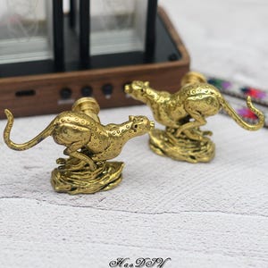 Könnte beinhalten: Zwei Messing-Gepardenfiguren in Laufpose. Die Geparden sind mit Flecken versehen und auf einem strukturierten Sockel montiert. Die Figuren werden auf einer hellen Oberfläche präsentiert. Der Etsy-Shop-Name und die URL stehen unten.