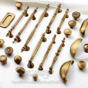 Op de afbeelding: Een assortiment antieke messing kastbeslagen, waaronder knoppen, grepen en handgrepen. De hardware heeft verschillende ontwerpen, van eenvoudige ronde knoppen tot sierlijke grepen met decoratieve details. De warme, gouden tinten van het messing creëren een klassieke esthetiek.