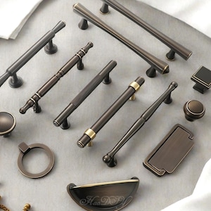 Puede incluir: Un surtido de herrajes para gabinetes en tonos bronce, que incluyen manijas, perillas y tiradores. La colección presenta varios diseños, desde tiradores de barra elegantes hasta perillas ornamentadas, con detalles dorados. El hardware está dispuesto sobre una superficie gris claro.