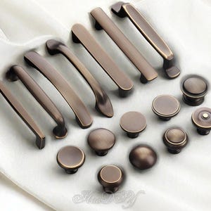 Könnte beinhalten: Eine Auswahl an bronzefarbener Schrankbeschlägen, darunter verschiedene Griffstile und runde Knöpfe. Die Griffe sind rechteckig und gebogen, während die Knöpfe flach und abgerundet sind. Die Beschläge werden auf einer weißen Oberfläche präsentiert.