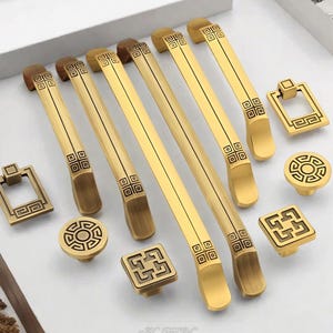 Op de afbeelding: Een verzameling goudkleurige kastbeslagen, waaronder lange stangtrekkers, vierkante trekkers en ronde knoppen. De hardware heeft een decoratief geometrisch patroon in een donkerdere tint. De stukken zijn gerangschikt op een wit oppervlak.