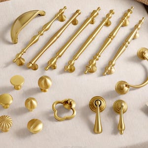 May include: A collection of gold-colored cabinet hardware, including various knobs, pulls, and handles. The hardware features ornate designs and a polished finish. The assortment includes different sizes and styles for furniture and cabinetry.