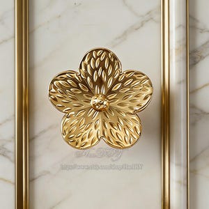 May include: A gold-coloured, flower-shaped cabinet knob. The knob is centered on a white marble surface with gold trim. The flower has detailed petal designs and a central circular accent.