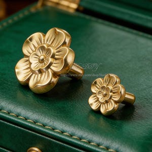 May include: Two gold-colored flower-shaped cabinet knobs. The knobs are made of metal and have a detailed floral design. The knobs are on a green surface. The Etsy shop URL is visible in the background.