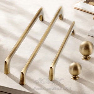 Könnte beinhalten: Ein Satz antiker Messing-Schrankbeschläge, darunter drei lange Stangengriffe und zwei runde Knöpfe. Die Beschläge haben eine warme, gealterte Oberfläche und sind auf einer hellen Oberfläche angeordnet. Die Griffe sind etwa 20 cm lang.