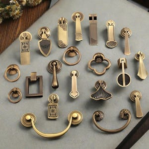 Può includere: Una collezione di maniglie e pomelli per armadi antichi in ottone di varie forme e dimensioni. L'hardware include maniglie ad anello, maniglie rettangolari e pomelli decorativi con disegni intricati. La tavolozza dei colori è principalmente oro e bronzo, con un'estetica vintage.