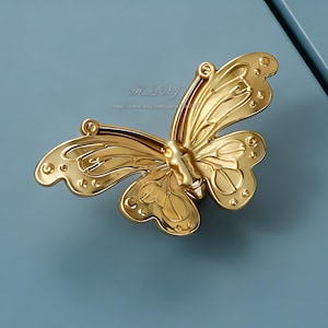 May include: A gold-coloured butterfly-shaped cabinet pull. The butterfly has detailed wing patterns and is mounted on a light blue surface. The hardware is made of metal and has a shiny finish. The Etsy shop name and URL are visible in the image.