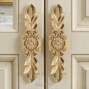 Puede incluir: Dos tiradores dorados ornamentados para armario con diseño floral y de hojas. Los tiradores están montados en una puerta de armario color crema. Los tiradores tienen un diseño floral central y están flanqueados por patrones de hojas simétricos.