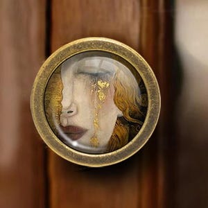 May include: A circular cabinet knob in antique brass, featuring a glass insert with a painting of a woman's face. The artwork depicts a woman with closed eyes and gold leaf details. Mounted on wood.