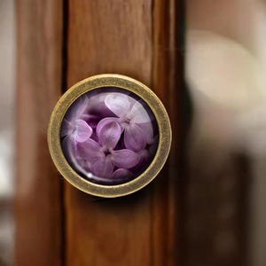 May include: A round cabinet knob with a brass finish and a glass insert. The insert features a close-up of purple lilac flowers. The knob is mounted on a dark brown wooden cabinet door.