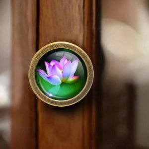 May include: A circular cabinet knob with a brass finish. It features a glass insert displaying a lotus flower design in pink, purple, and blue hues set against a green backdrop. The knob is attached to a wooden cabinet door.