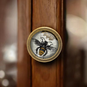 以下が含まれることがあります： ハニカムに描かれたビンテージの蜂のイラストが施された、ガラス製のインサートが付いた丸い真鍮製のキャビネットノブです。