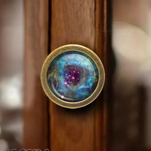 Könnte beinhalten: Ein Messing-Schrankknauf mit einem runden Glaseinsatz, der ein farbenfrohes Nebelbild zeigt. Der Nebel ist ein wirbelnder Mix aus Blau, Lila und Rosa mit einer Streuung von weißen Sternen.