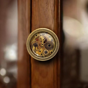 以下が含まれることがあります： 時計仕掛けのディテールが見える透明なガラス製のインサートが付いた、真鍮色の丸いキャビネットノブです。