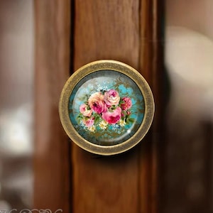 May include: A round, antique gold-toned cabinet knob with a glass insert featuring a floral design of pink roses and white daisies on a blue and gold patterned background.