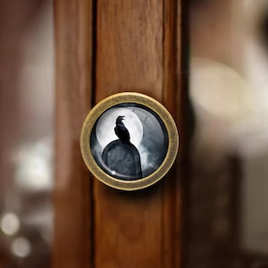 May include: A round, gold-toned cabinet knob with a glass insert featuring a black raven perched on a tombstone in front of a full moon.