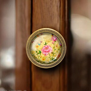 May include: A round, antique gold cabinet knob with a floral design. The knob features a pink flower with yellow and green leaves and butterflies on a cream background.