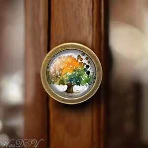 May include: A round, brass cabinet knob with a glass insert featuring a watercolor painting of a tree with green, orange, and yellow leaves.