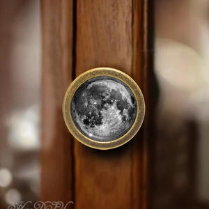 May include: A round, brass cabinet knob with a full moon image in the center. The moon is a black and white photo with a textured surface.