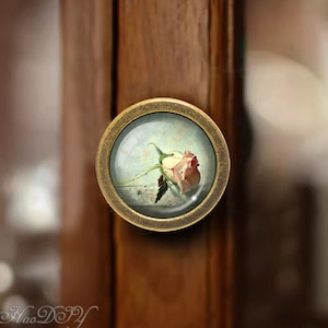 Könnte beinhalten: Ein runder, messingfarbener Schrankknopf mit einem Glaseinsatz, der ein gemaltes Bild einer einzelnen, rosafarbenen Rose zeigt.