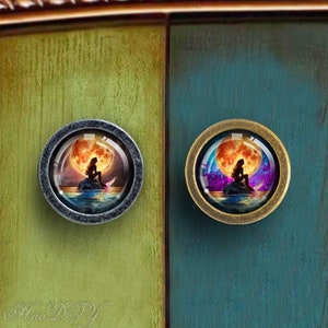 May include: Two round cabinet knobs featuring a mermaid sitting on a crescent moon. The knob on the left has a silver finish and the knob on the right has a brass finish.
