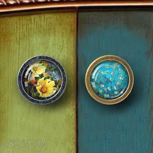 May include: Two round cabinet knobs with floral designs. The left knob has a gray metal base and a glass insert with a yellow flower design. The right knob has a brass base and a glass insert with a blue and white floral design.