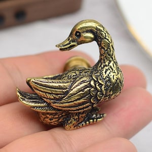 May include: A detailed brass duck figurine with intricate feather detailing. The duck has a curved neck and is posed in a resting position. The figurine is a warm gold color and appears to be made of metal. The Etsy shop name and URL are visible at the bottom of the image.