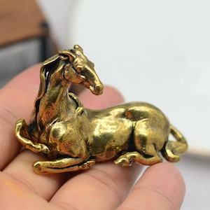 May include: A brass figurine of a horse in a reclining position. The sculpture is a warm gold colour, with detailed features of the horse's mane, body, and legs. The horse is lying down, with its head raised.