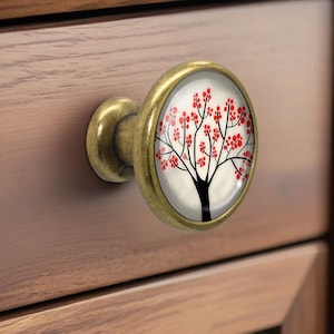 May include: A decorative cabinet knob with a brass finish. The knob features a round, glass-covered design with a black tree and red blossoms against a white background. The knob is attached to a wooden drawer.