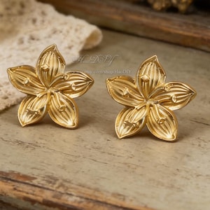 May include: A pair of gold-colored flower-shaped earrings. Each earring features five petals with detailed veining. The earrings are displayed on a light-colored wooden surface, with a blurred background including a mirror and a glass bottle.