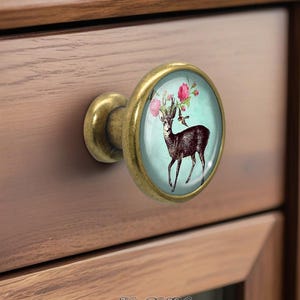 Può includere: Pomello rotondo per armadio con finitura in ottone. Il pomello presenta un inserto in vetro con un'illustrazione di cervo adornata da fiori rosa e un uccello su sfondo turchese. Il pomello è attaccato a un mobile in legno.