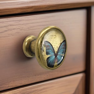 May include: A cabinet knob with a brass finish. The knob has a round glass insert with a blue butterfly design on a vintage paper background. It is attached to a wooden drawer. The knob is made of brass.